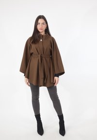 Mantello di lana marrone con cintura, caratterizzato da un colletto alto e un singolo bottone metallico. Abbinato a jeans skinny grigi e stivaletti neri.