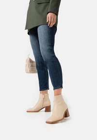 Beżowe skórzane botki za kostkę z kwadratowym noskiem, zamkiem bocznym i słupkowym obcasem, połączone z ciemnoniebieskimi dżinsami i jasną torebką.