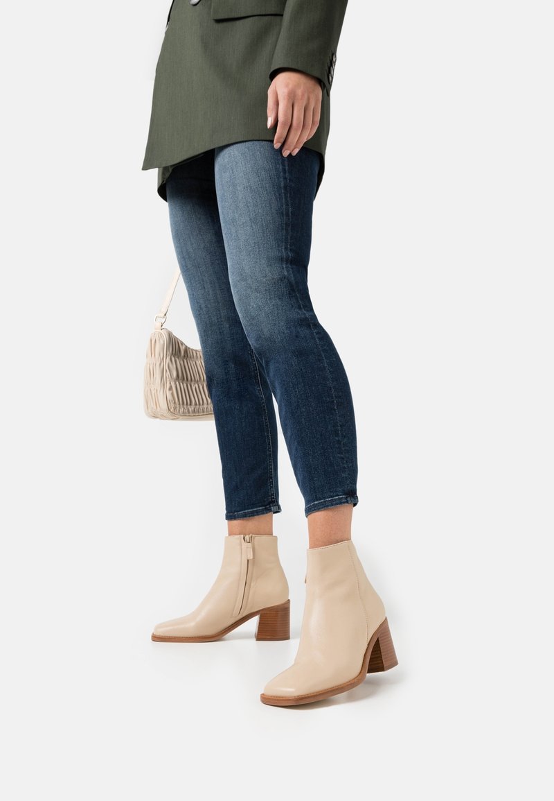 Beżowe skórzane botki za kostkę z kwadratowym noskiem, zamkiem bocznym i słupkowym obcasem, połączone z ciemnoniebieskimi dżinsami i jasną torebką.
