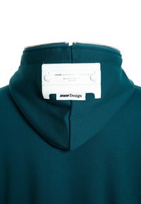 Sudadera teal con cuello con cremallera. Presenta una etiqueta de cuero blanca con el nombre de la marca y detalles de estilo. Tejido suave y diseño estructurado.