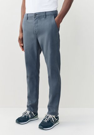 Homme portant un pantalon gris ajusté, un t-shirt blanc et des baskets noires avec des lacets blancs, debout avec une main dans la poche.