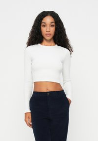 Biała, długa bluzka crop top z prążkowanego materiału, zestawiona z granatowymi spodniami z detalem guzika i bocznymi kieszeniami.