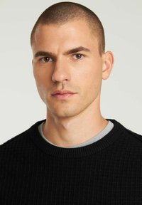 Maglione nero in maglia con texture, colletto a girocollo a costine, indossato sopra una t-shirt grigia. Primo piano del viso e delle spalle del modello.