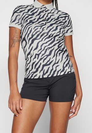 Kvinde iført en kortærmet zebra-stribet polo og sorte shorts, der viser synlige tatoveringer på begge arme mod en ensfarvet baggrund.