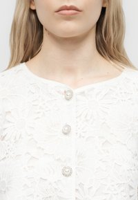 Naise kael ja ülaosa rinna juures, kandes valget lillelist pitspluusi pärli- ja kivikestega nuppudega, heledat blondi juukseid õlgadel nähtaval.