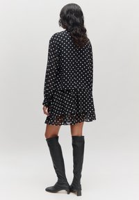 Schwarzes Polka-Dot-Kleid mit langen Ärmeln, gerüschtem Saum und durchscheinendem Stoff, kombiniert mit kniehohen schwarzen Stiefeln. Ansicht von hinten.