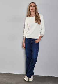 Weißes, strukturiertes Langarmoberteil mit Rundhalsausschnitt, kombiniert mit dunkelblauen, geraden Jeans und hellen Sneakern, vor einem grauen Hintergrund.