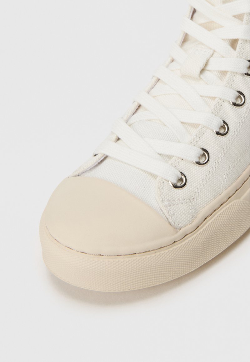 Weißes Canvas-Hochtop-Sneaker mit beigefarbenem Gummi-Zehenkappe, silbernen Ösen und flachen weißen Schnürsenkeln, mit einer strukturierten Stoffoberfläche.