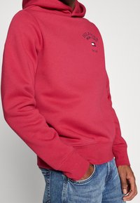 Czerwona bluza z kapturem, z kieszenią z przodu, regulowanym sznurkiem oraz haftowanym logo. Fakturowana tkanina, dopasowane mankiety i dół. Noszona z niebieskimi dżinsami.