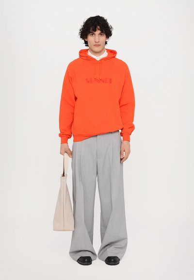 Orangene Kapuzenjacke mit "SUNNEI"-Logo, kombiniert mit hellgrauen, weiten Hosen und schwarzen Schuhen. Die Person hält eine beige Tragetasche.