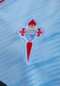 Una tela azul clara presenta un escudo bordado con un diseño en rojo y blanco, que incluye elementos de una corona y una flor de lis.