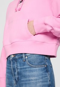 Felpa di cotone rosa con tasca frontale, chiusura con cordino e polsini a coste, abbinata a jeans di denim blu chiaro con bottone e tasca.
