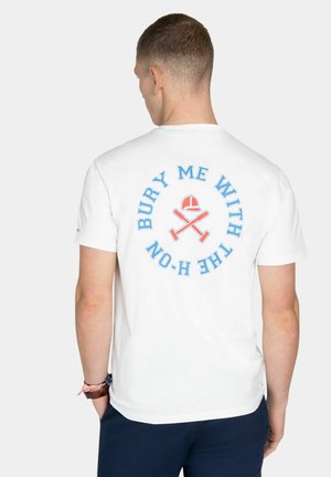 Hombre con camiseta blanca con texto azul "ENTIÉRRAME CON LA H-O-N" y bate de cricket rojo cruzado y casco en la espalda.