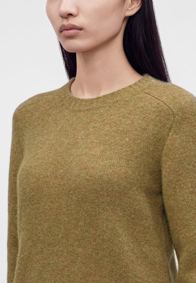 Femme portant un pull en laine vert mousse à manches longues avec un col rond côtelé, visible des épaules jusqu'au milieu de la poitrine.