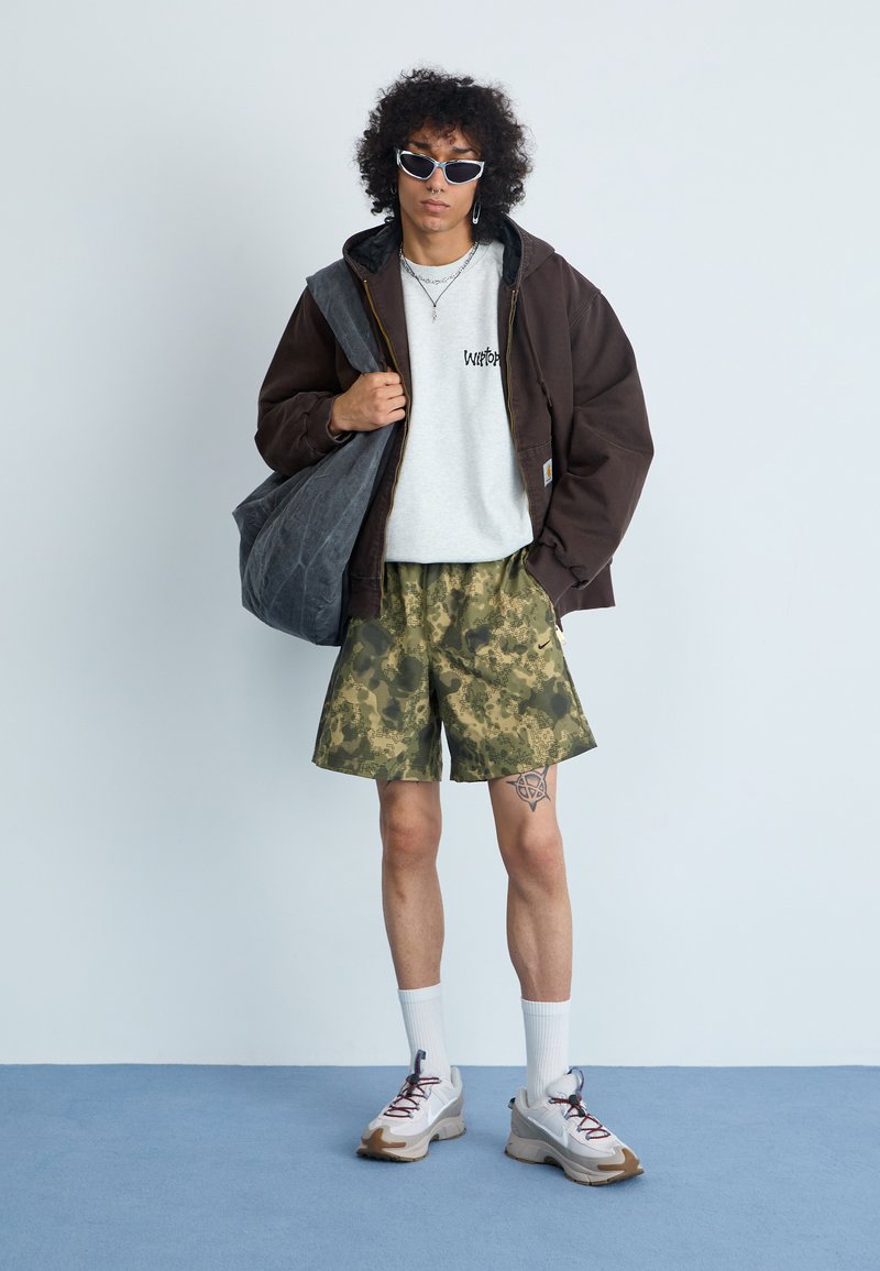 Brun huva jacka, grå grafisk t-shirt, kamouflage shorts, vita strumpor, grå och bruna sneakers, silveraccessoarer, lockigt hår.