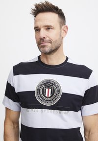 Svītrainā īsām piedurkņu T-kreklā tumši zilā un baltā krāsā ir apļveida logo ar lauru akcentiem un teksts "1924 SPORTSWEAR" apakšā.