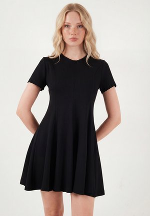 Noor naine blondide juustega, kes kannab lühikeste varrukatega musta kleiti, seisab käed selja taga tavalise valge tausta ees.