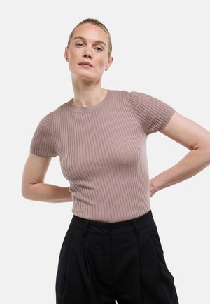 Femme aux cheveux blonds portant un haut mauve en maille côtelée à manches courtes et un pantalon noir taille haute, posant avec les mains derrière le dos.