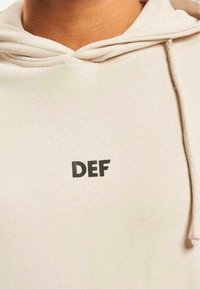 Lys beige hettegenser med svart "DEF" trykk foran. Laget av myk stoff med snøring i hetta og ribbestrikkede mansjetter.