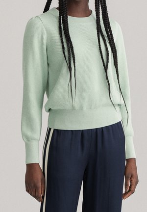 Personne portant un pull côtelé vert clair et un pantalon bleu marine avec des bandes blanches sur les côtés, debout devant un fond uni.