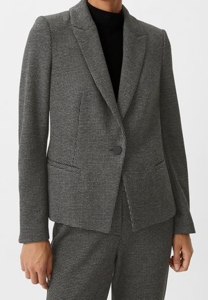 Frau trägt eine taillierte graue Hahnentritt-Blazer mit einem Knopf und passenden Hosen, kombiniert mit einem schwarzen Top darunter.