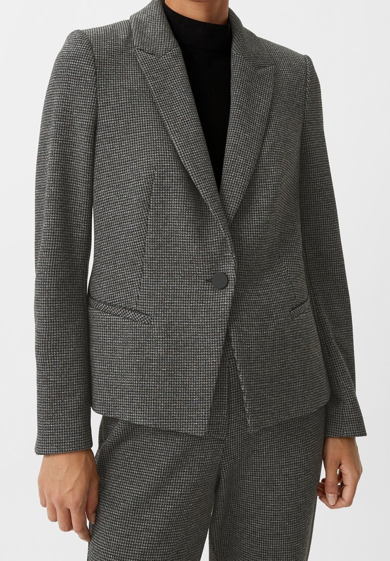 Femme portant un blazer gris pied-de-poule ajusté avec un bouton et un pantalon assorti, associé à un top noir en dessous.