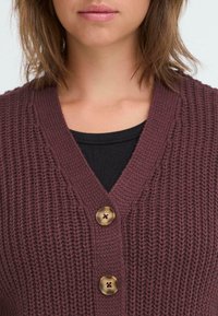 Neulepusero syvässä marooninvärissä, jossa on V-pääntie, kaksi messinkiä nappia ja teksturoitu ribbikuvio. Musta toppi alla.