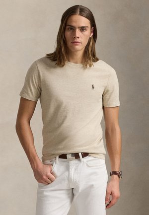 Ung mand med langt hår iført en beige kortærmet skjorte og hvide bukser, stående med den ene hånd i lommen mod en ensfarvet baggrund.