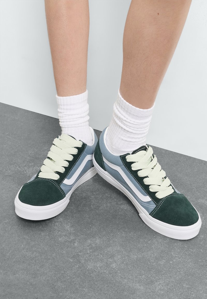 Baskets en daim vert et bleu avec des accents blancs, lacets ronds, design à coupe basse et semelle en caoutchouc texturée. Chaussettes blanches côtelées visibles.