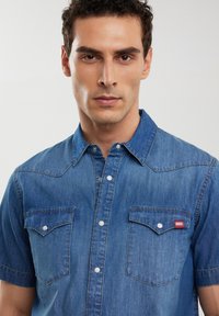 Camicia in denim a maniche corte di un blu medio. Presenta due tasche sul petto, bottoni a pressione, colletto a punta e un piccolo logo rosso sul taschino.