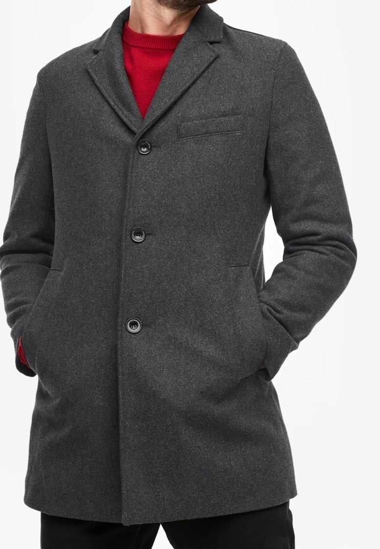 Uomo che indossa un cappotto di lana grigio scuro con bottoni sopra un maglione rosso, mani nelle tasche, rivolto leggermente a sinistra con la testa parzialmente visibile.