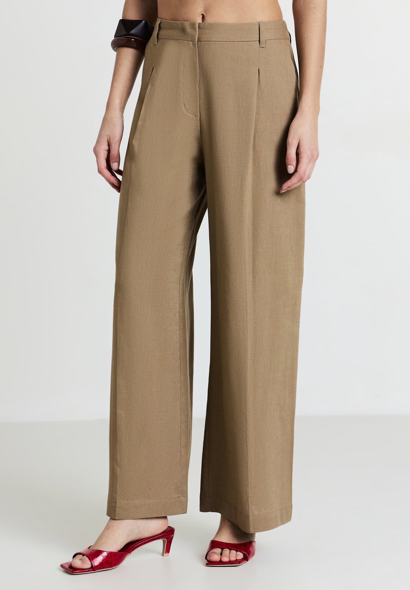 Pantaloni a gamba larga in un tessuto marrone chiaro, caratterizzati da pieghe, una vestibilità su misura e una texture liscia, abbinati a mule con tacco rosso lucido.