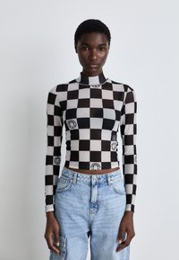 Long-sleeve top in black and white checkerboard pattern with graphics, featuring a mock neck and a fitted design, paired with light blue jeans.
