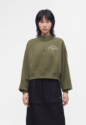 Jeune personne aux cheveux noirs raides porte un sweat-shirt Nike vert olive et une jupe noire avec des poches sur un fond blanc uni.