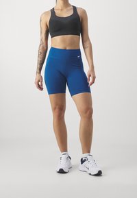 Zwarte geribbelde sportbeha, blauwe hoge taille fietsbroek en witte sportschoenen. Opvallend Nike-logo op de broek. Minimalistisch ontwerp.