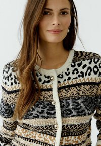 Knopfburgunder in Strickstoff mit verschiedenen Mustern in Schwarz, Creme und Beige. Goldene Knopfdetails runden das Design ab.