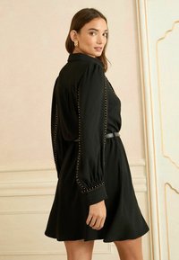 Rochie neagră cu mâneci bufante, talie ajustată și fustă evazată. Detalii din metal aurit pe mâneci. Textură de țesătură netedă.