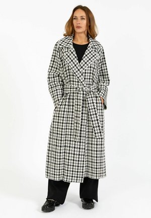 Nő fekete-fehér kockás, hosszú, öves kabátban, fekete nadrágban és fekete cipőben, kezét a kabát zsebében tartva áll.