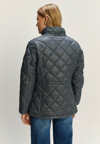 Žena s blond vlasmi, oblečená v prešívanej tmavosivej bunde a modrých džínsach, otočená chrbtom k jednému svetlému pozadiu.