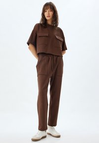 Bruine cropped top met twee voorzakken, gecombineerd met bijpassende tapered pants met een elastische tailleband en zijzakken. Witte sneakers.