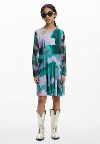 Langärmliges Kleid mit mehrfarbigem Aquarellmuster in Grün, Rosa und Lila, mit ausgestelltem Rock. Kombiniert mit silbernen Cowboy-Stiefeln.