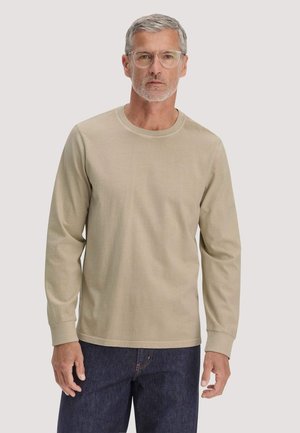 Mann mit grauen Haaren und Bart, der klare Brillen, ein beige langärmeliges Hemd und dunkelblaue Jeans trägt, stehend vor einem einfarbigen Hintergrund.