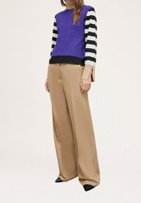 Pull en maille violet avec des manches rayées noir et blanc, associé à un pantalon beige à jambes larges. La tenue se caractérise par des lignes épurées et un design minimaliste.