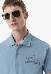 Jasnoniebieska koszulka polo z teksturowanej tkaniny, z podwójnym zapięciem na guziki i szarym logo krokodyla na piersi.