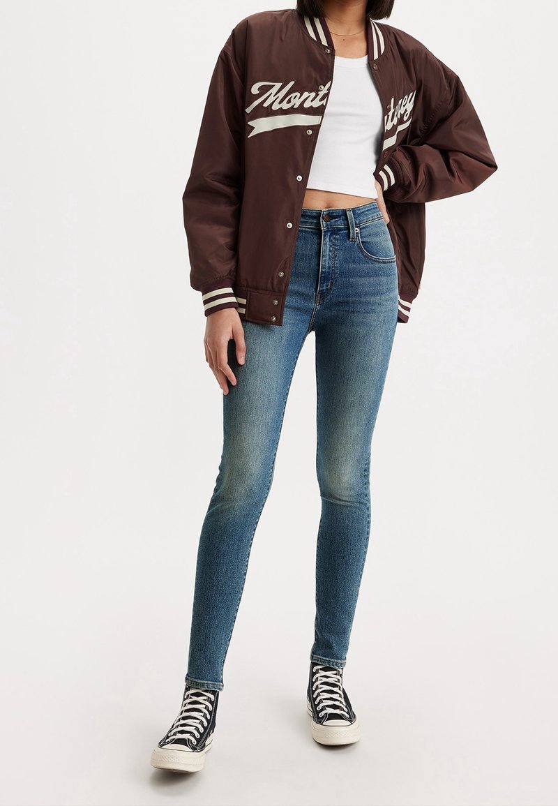 Chaqueta varsity marrón con letras blancas, llevada sobre una camiseta corta blanca. Jeans ajustados de mezclilla azul claro, zapatillas de lona altas negras con suelas blancas.