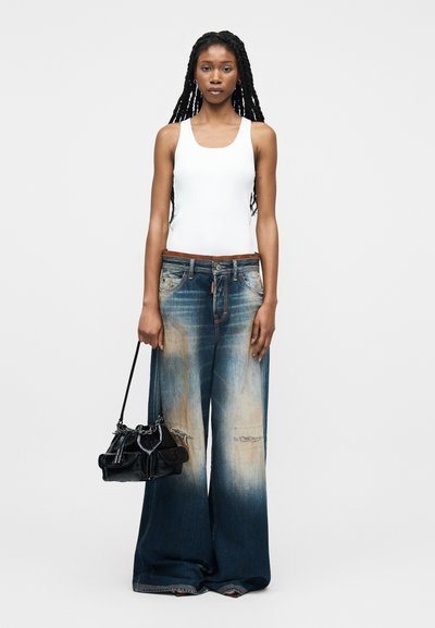 Γυναίκα με μακριά πλεξουδωτά μαλλιά, ντυμένη με λευκό αμάνικο τοπ και φαρδιά ξεθωριασμένα τζιν, κρατώντας μια μαύρη χειραποσκευή σε φόντο απλό και φωτεινό.