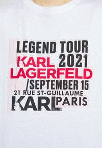 Bílé bavlněné tričko s výrazným, vícebarevným nápisem. Klíčové detaily zahrnují "LEGEND TOUR," "KARL LAGERFELD" a adresu v Paříži.