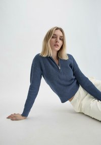 Blauwe gebreide trui met een half-zip kraag, geribbelde textuur en lange mouwen. Gecombineerd met lichtgekleurde broeken, voor een effen achtergrond.