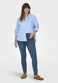 Modrá a biela pruhovaná košeľa s gombíkmi a vyrolovanými rukávmi, kombinovaná s tmavými džínsami a svetlohnedými nazúvacími topánkami. Minimalistický dizajn, voľný strih.