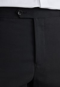 Gros plan sur la ceinture d’un pantalon noir avec une fermeture par bouton, porté avec une chemise blanche rentrée.